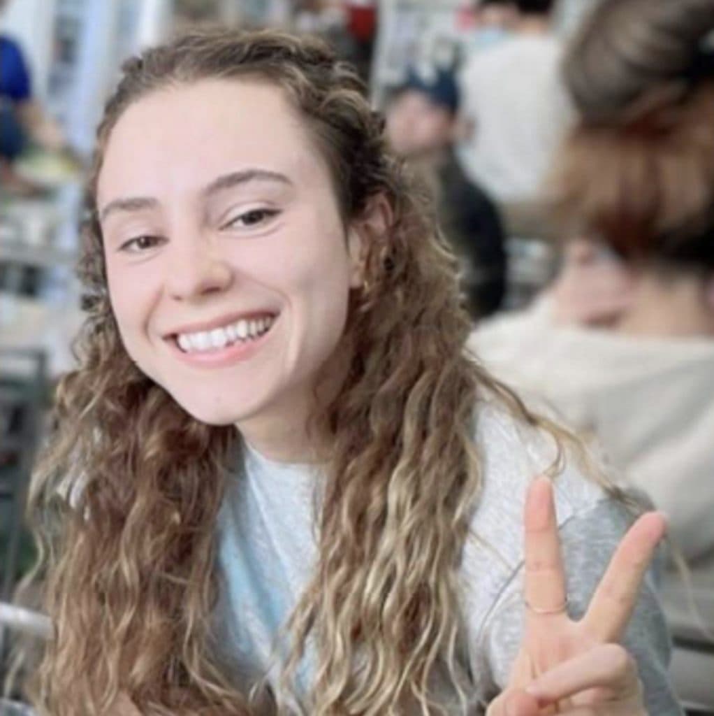 A smiling young woman with long curly hair making the peace sign.